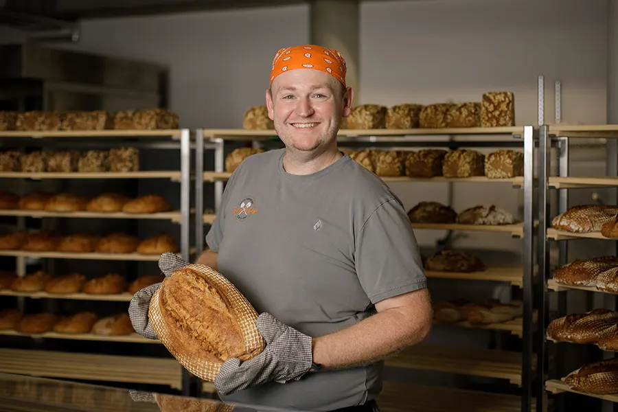 David von der Schöne Brotmanufaktur hält ein frisch gebackenes Brot vor den Brotregalen seiner Backstube.